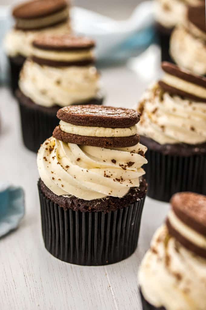 A chocolate cupcake with white frosting and a cookie sitting on top. It is surrounded by more cupcakes.