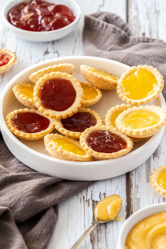 A white platter filled with jam tarts on a dark grey tea towel. It sits on a white wooden background