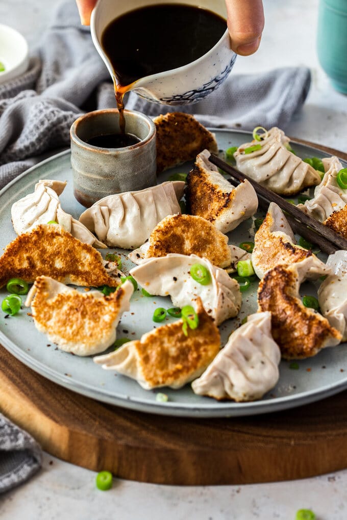A plate of 16 pork gyoza. The grey plate sits on a wooden platter. Sauce is being poured into a grey pot of behind them.