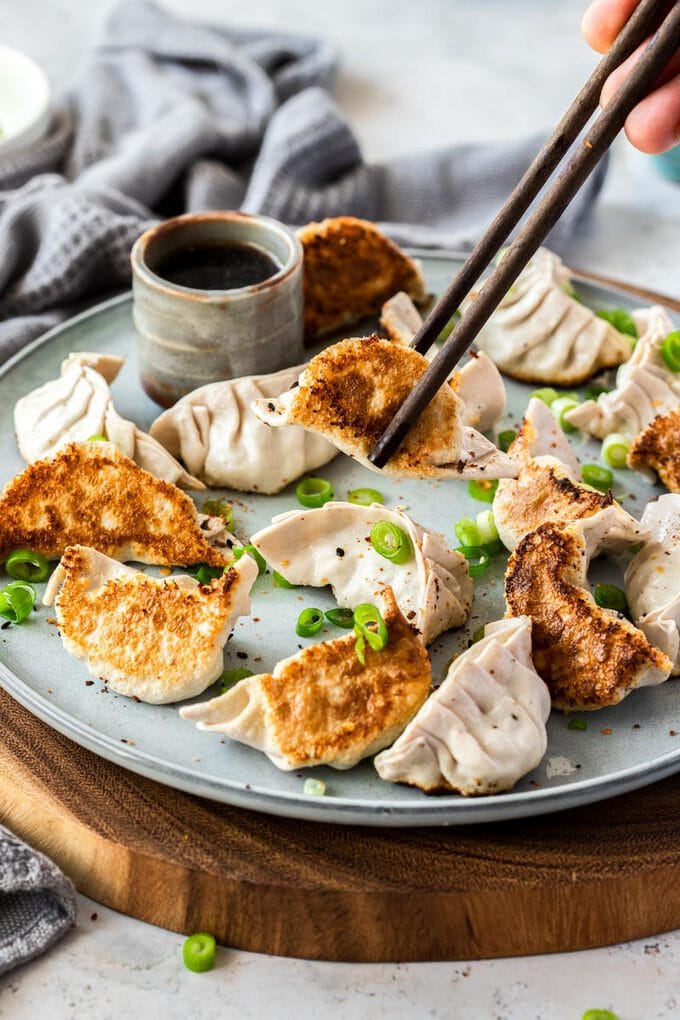 16 pork gyoza on a grey plate with chopsticks picking one up. A small grey pot of sauce behind