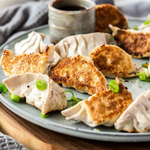 10 pork gyoza on a grey plate on a wooden platter. A small grey pot of dipping sauce behind them.