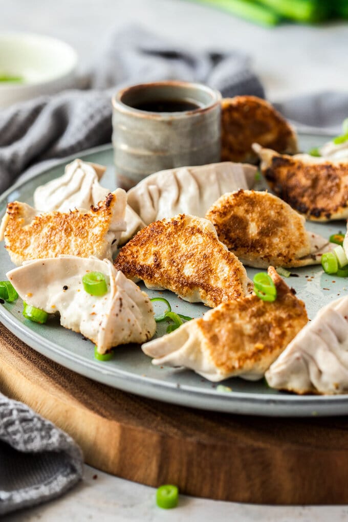 10 pork gyoza on a grey plate, that sits on a wooden platter. A small grey pot of sauce sits behind them