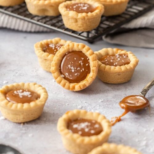 A group of caramel cookie cups on a grey surface. A spoon filled with caramel next to them and a rack of more cookies behind
