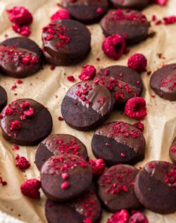 A batch of dark chocolate shortbread cookies, sprinkled with freeze-dried raspberries on a sheet of brown baking paper