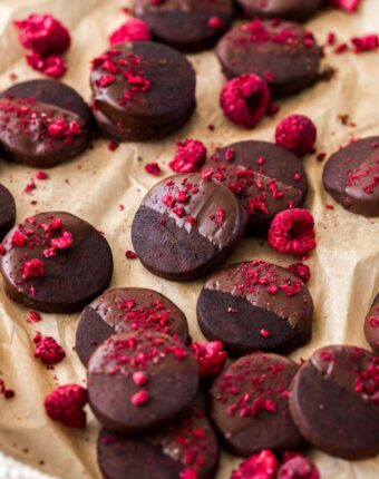 A batch of dark chocolate shortbread cookies, sprinkled with freeze-dried raspberries on a sheet of brown baking paper