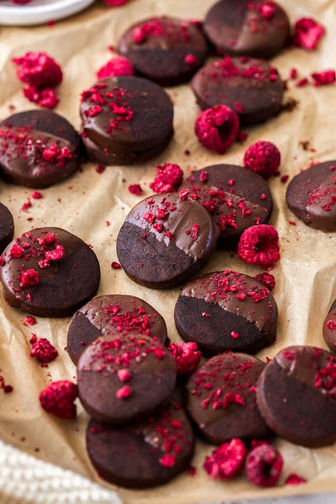 A batch of dark chocolate shortbraed cookies, sprinkled with freeze-dried raspberries on a sheet of brown baking paper