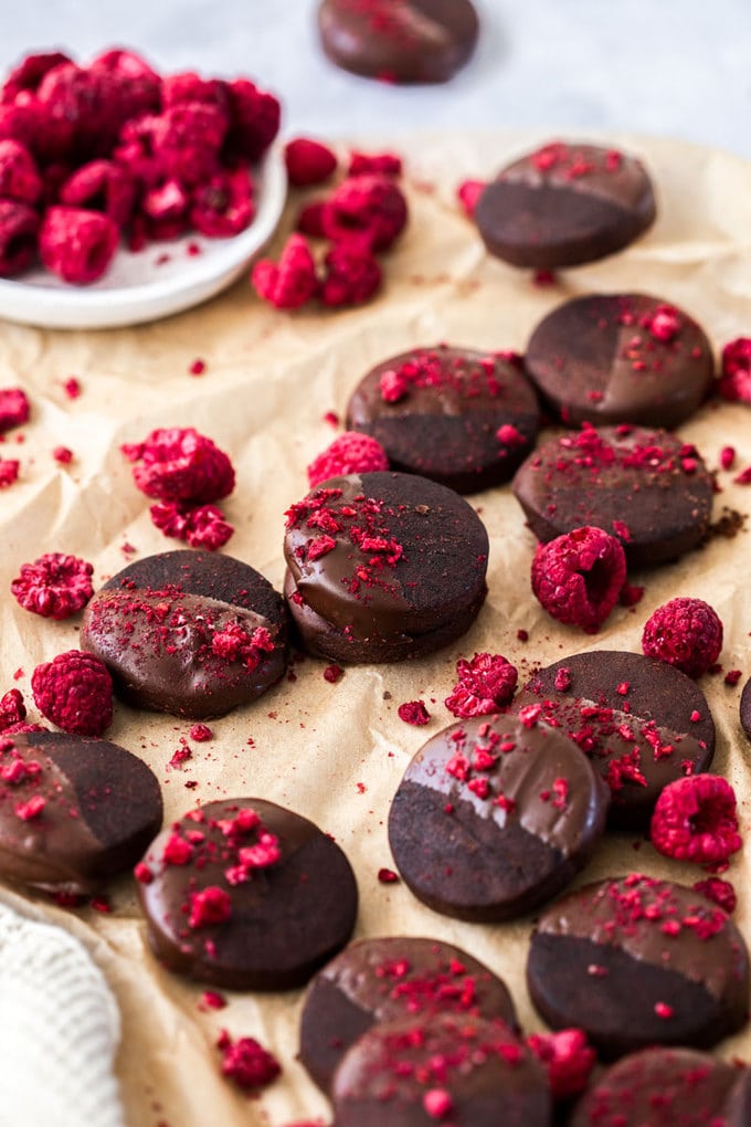 Dark chocolate shortbread cookies, sprinkled with freeze-dried raspberries laying on a sheet of brown baking paper, a plate of freeze dried raspberries in the back