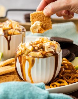 A cookie that's been dipped into a white bowl full of snickers dip. Surrounded by more cookies and pretzels
