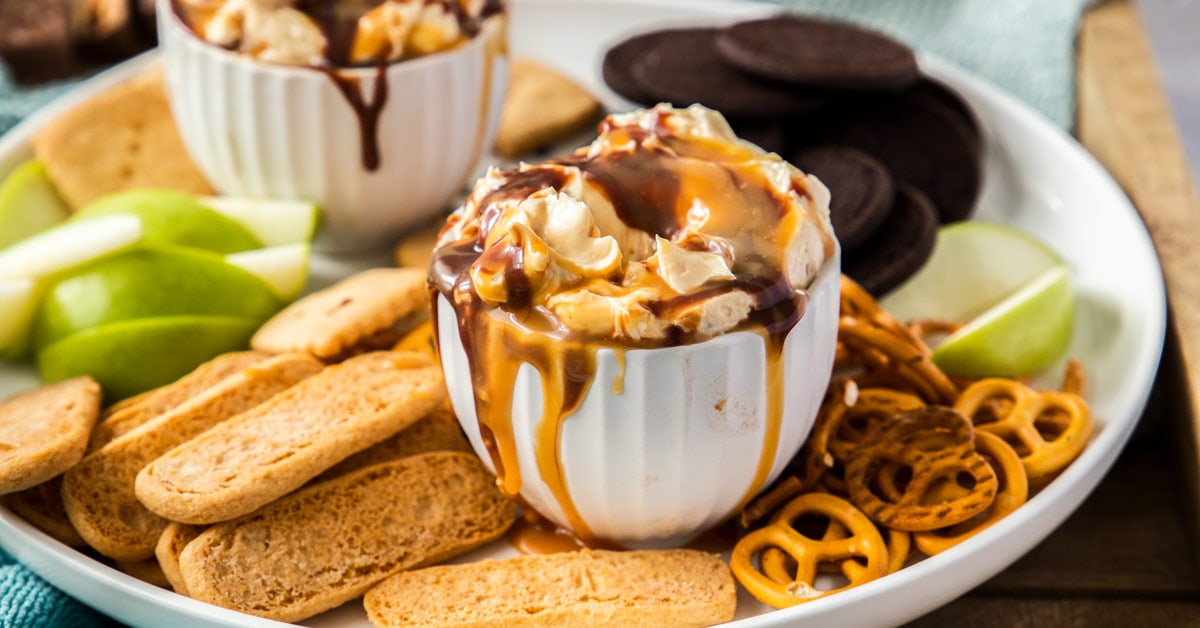 A white bowl filled with cheesecake dip with chocolate and caramel dripping down the side. It's surrounded by cookies and pretzels
