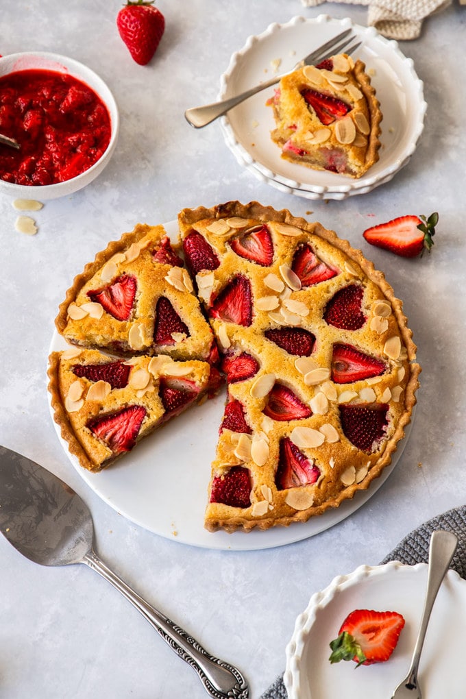 birdseye view of a strawberry tart on a white plate. A cake slice sits in front and dessert plates are set around it