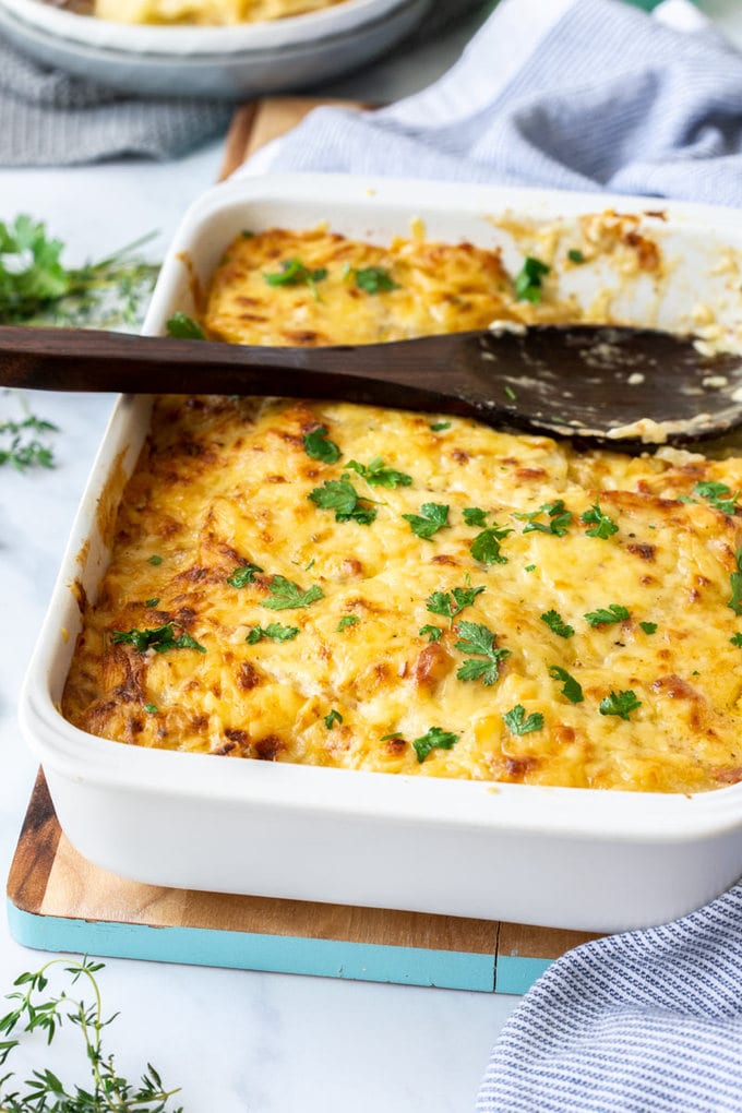 A white casserole dish filled with cheesy scalloped potatoes with a wooden spoon sitting on top.