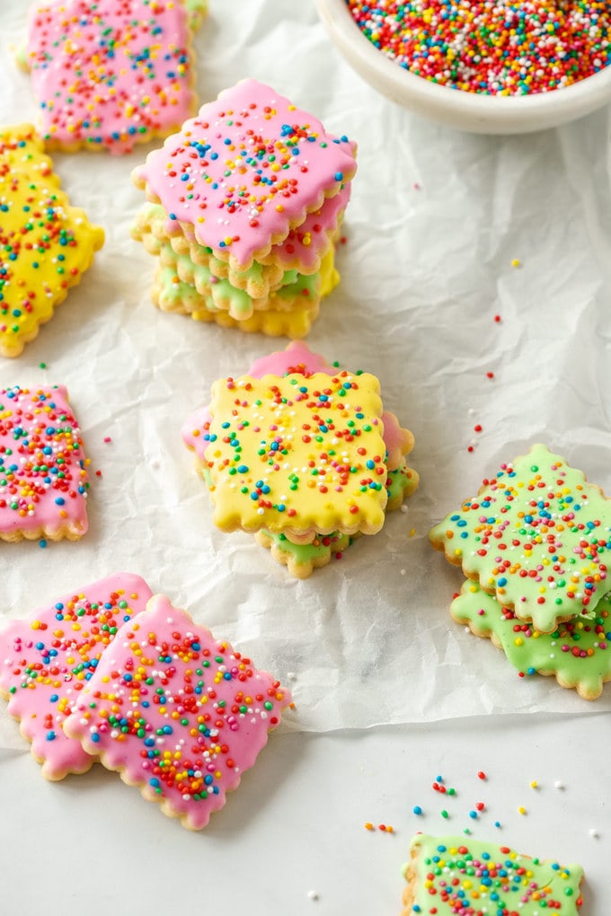 Iced cookies covered in sprinkles on a sheet of baking paper next to a bowl of sprinkles.