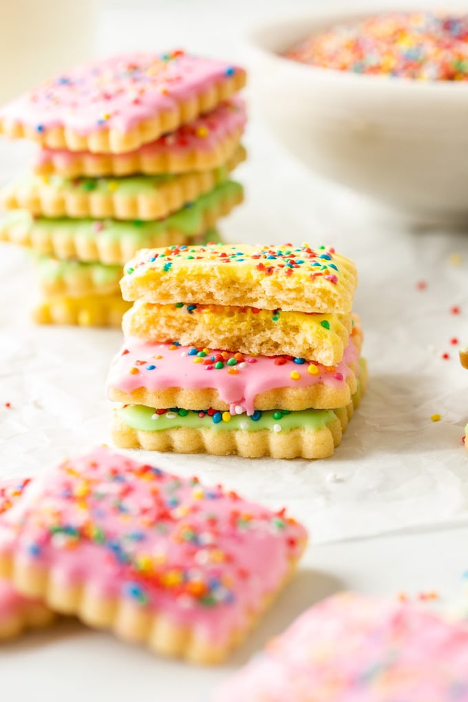 Stacks of coloured iced cookies covered in sprinkles on a sheet of baking paper.