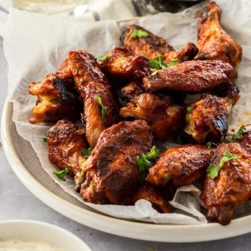 BBQ chicken wings on baking paper on a grey plate. A bowl of ranch dip sits in front and avocado dip sits behind.