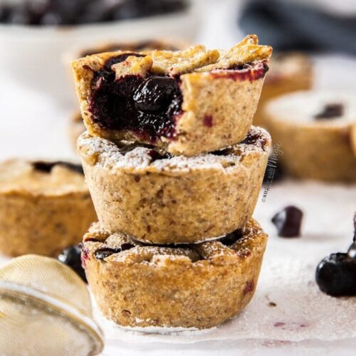 A stack of 3 mini blueberry pies with more surrounding it. A white bowl full of blueberries sits in the background