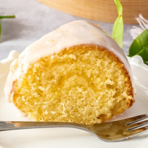A slice of vanilla butter cake on a white cake plate with a fork in front. Some lemon tree leaves sit around it