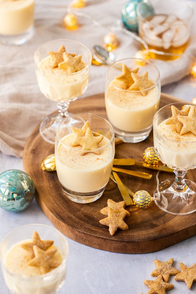Glasses filled with eggnog panna cotta on a wooden board, surrounded by shortbread and festive ornaments.