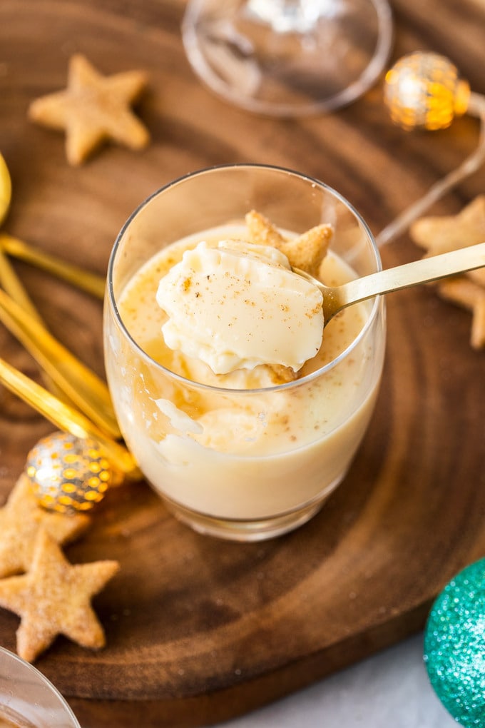 A spoon scooping eggnog panna cotta out of a glass. It sits on a wooden board surrounded by shortbread and festive ornaments