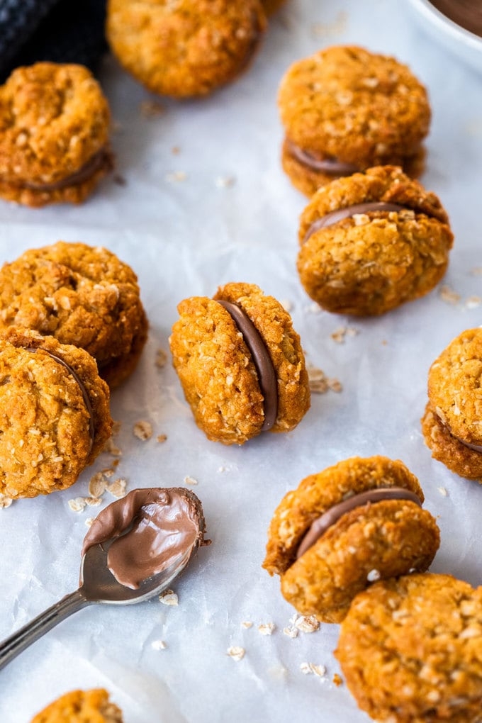 A batch of chocolate filled oatmeal cookies lying on a sheet of baking paper with a spoon covered in chocolate next to them