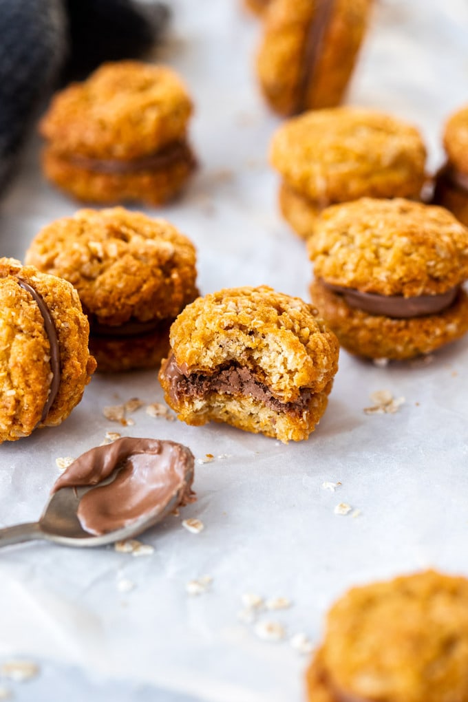 A batch of chocolate filled oatmeal cookies lying on a sheet of baking paper with a spoon covered in chocolate next to them. One has a bite taken out of it
