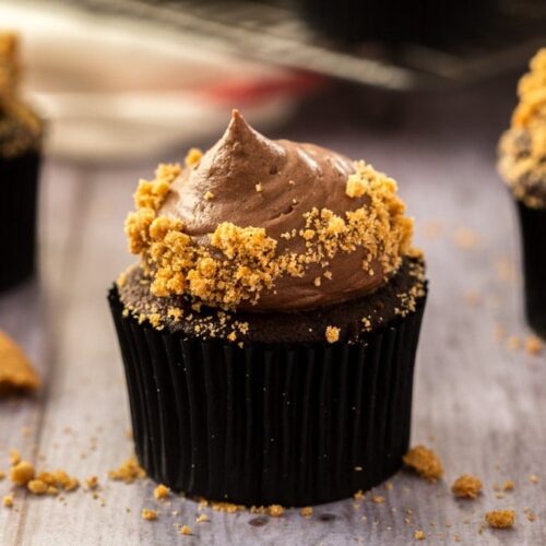 Closeup of a chocolate cupcake with chocolate frosting and covered in cookie crumbs on a grey wood surface