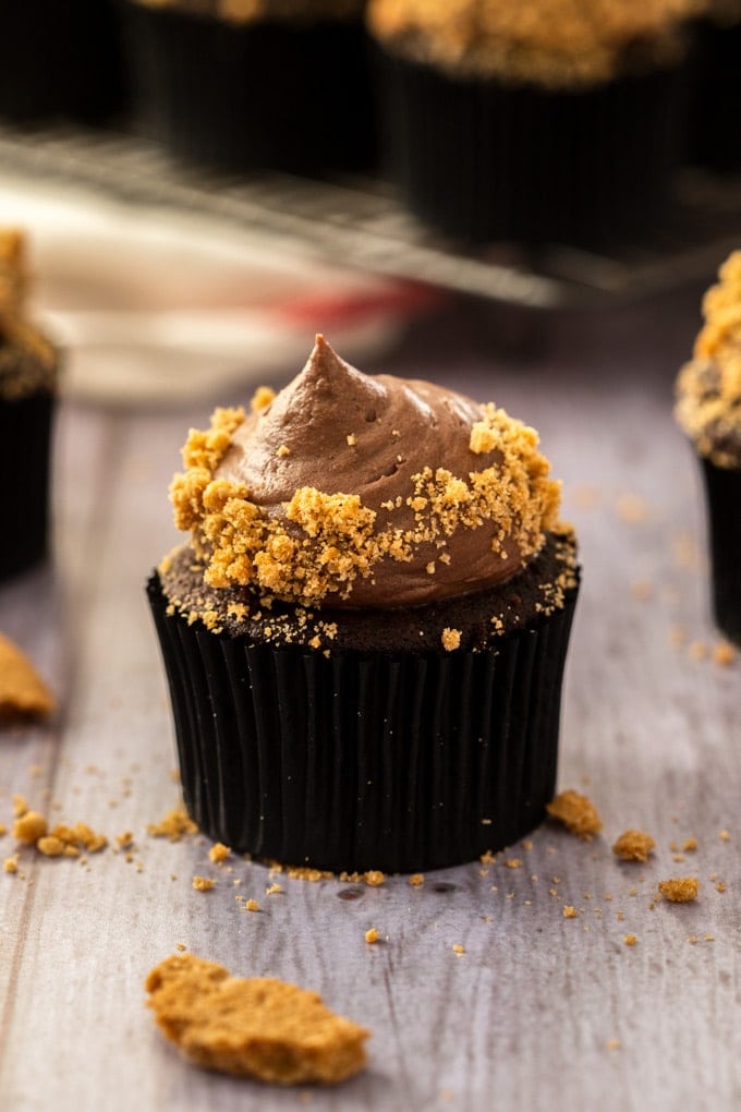 Closeup of a chocolate cupcake with chocolate frosting and covered in cookie crumbs on a grey wood surface.