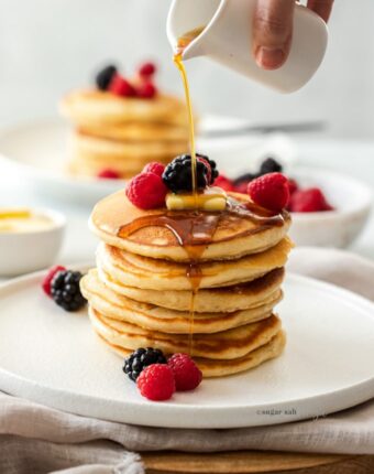 A stack of pancakes on a white plate with berries and maple syrup being poured over them.