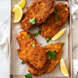 Chicken schnitzels and wedges of lemon on a gold baking tray with a bowl of green salad leaves in the background.