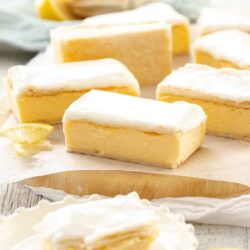 A batch of vanilla custard slice on a sheet of baking paper on a wooden board. Once sits on a plate in front with a bite taken out