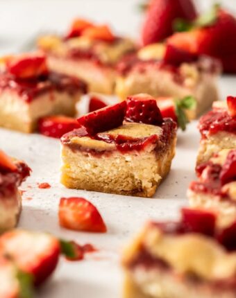 A cheesecake bar topped with strawberry topping sitting on a white background surrounded by strawberries and more bars