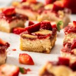 A cheesecake bar topped with strawberry topping sitting on a white background surrounded by strawberries and more bars