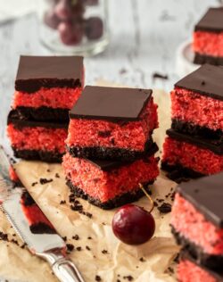 Stacks of cherry ripe slice on brown parchment paper, with a knife in front and a glass of cherries as the back
