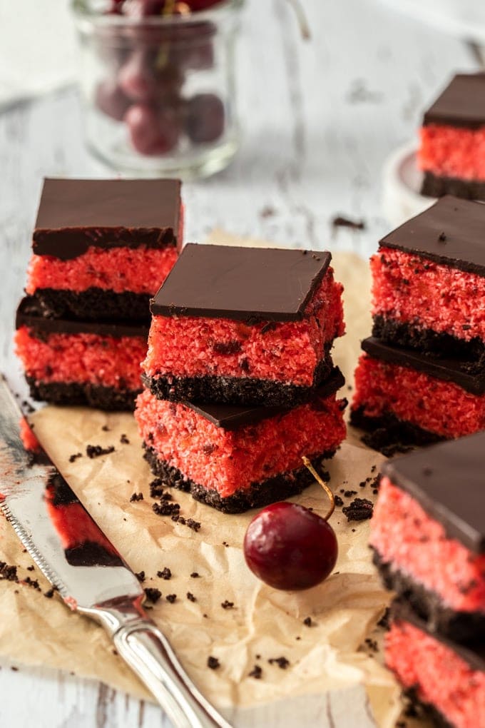 Stacks of cherry ripe slice on brown parchment paper, with a knife in front and a glass of cherries as the back