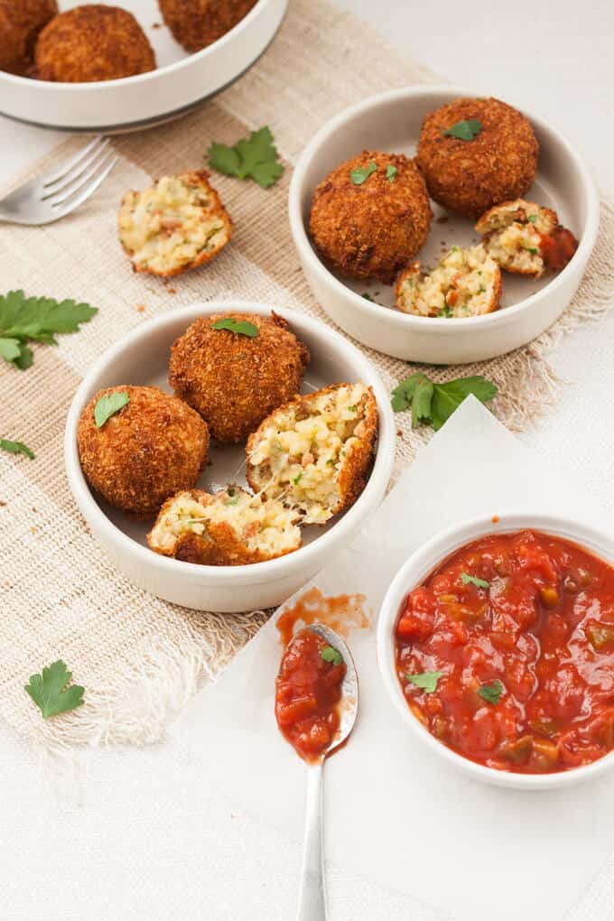 Two small dishes filled with arancini and a small pot of marinara sauce.