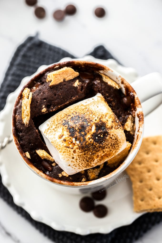 A white cup on a saucer filled with chocolate cake and toasted marshmallow. It sits on a grey crocheted napkin