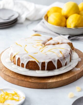 A lemon cake drizzled with white icing on a marble platter and wooden board. Lemons in the background.