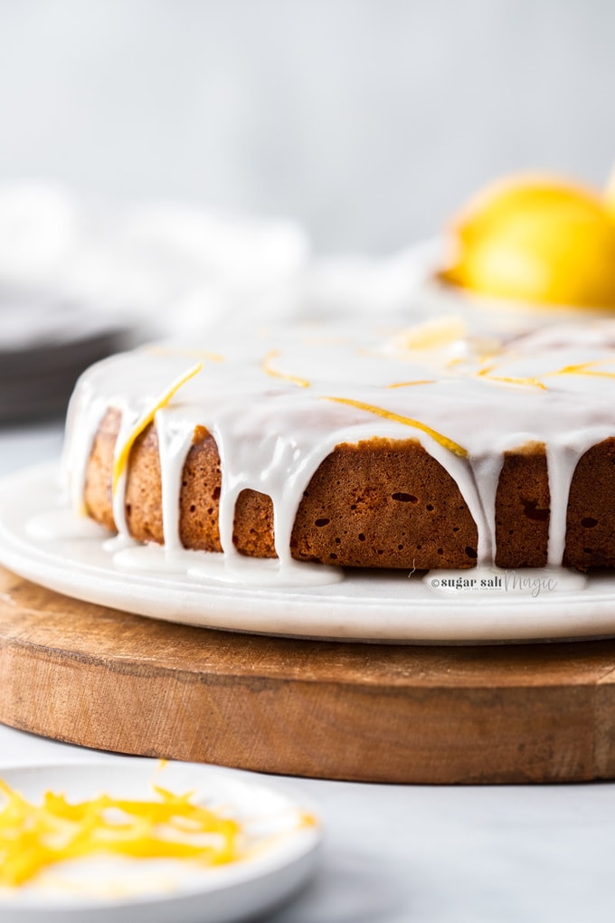 A lemon cake drizzled with white icing on a marble platter and wooden board. Lemons in the background.