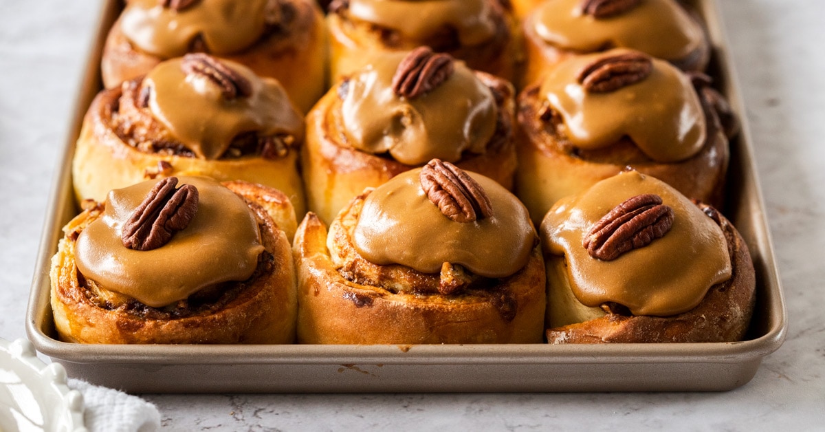 A batch of baked cinnamon rolls topped with coffee icing and pecans on a gold baking tray.