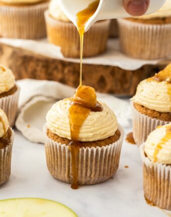 An apple pie cupcake being drizzled with maple syrup, surrounded by more cupcakes and apple slices, on a white background