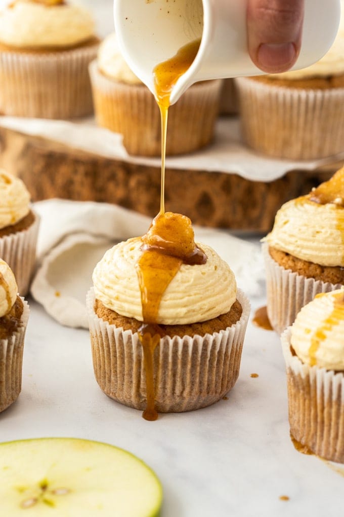 An apple pie cupcake being drizzled with maple syrup, surrounded by more cupcakes and apple slices, on a white background