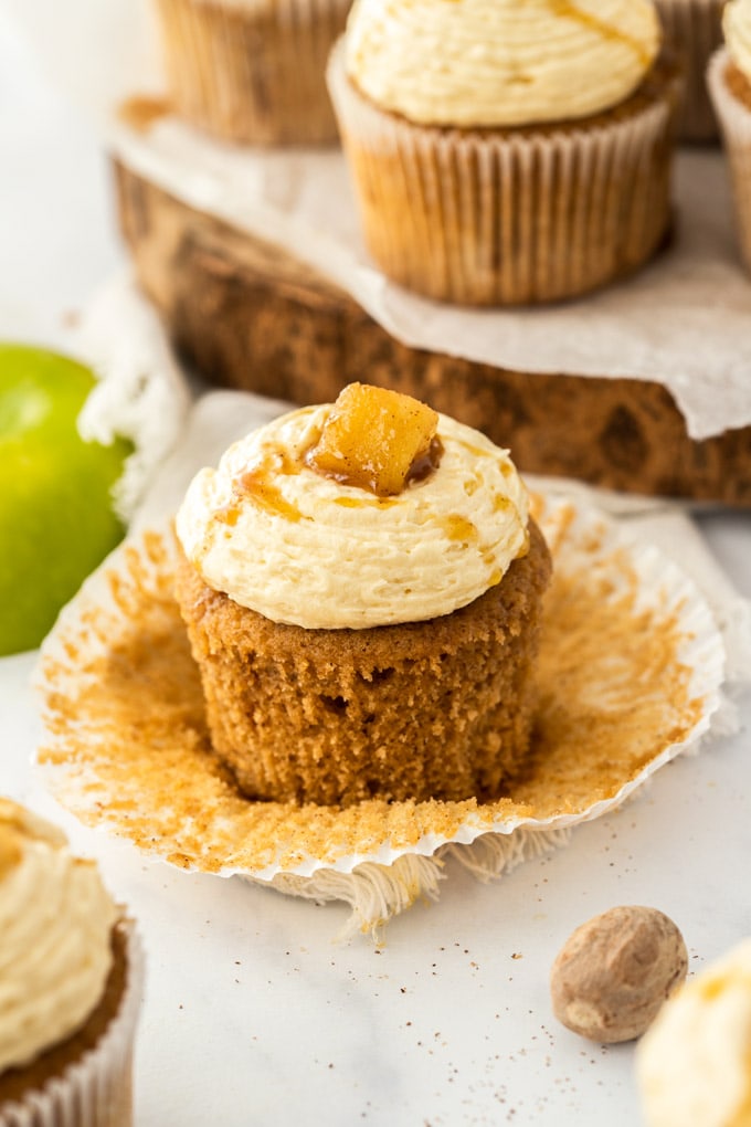 A closeup of an apple pie cupcake with the paper peeled away showing the texture of the cake. It sits on a marble worktop