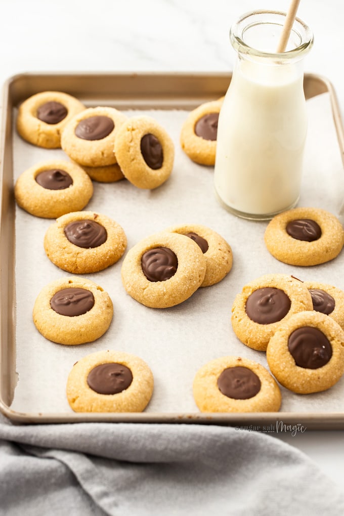 A batch of cookies with chocolate centres on a sheet of baking paper in a baking tin. A small bottle of milk at the back
