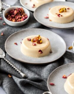 A creamy panna cotta dessert on a grey plate, topped with pistachio toffee and rose petals