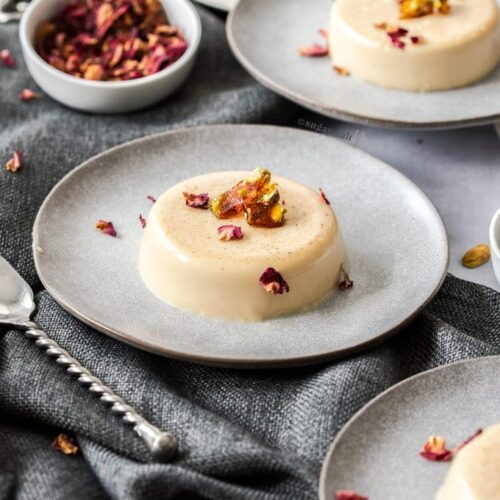 A creamy panna cotta dessert on a grey plate, topped with pistachio toffee and rose petals