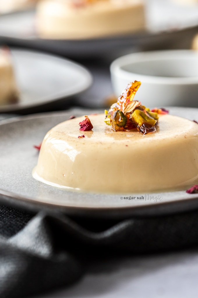 Closeup of a panna cotta on a grey plate, topped with toffee pieces and rose petals