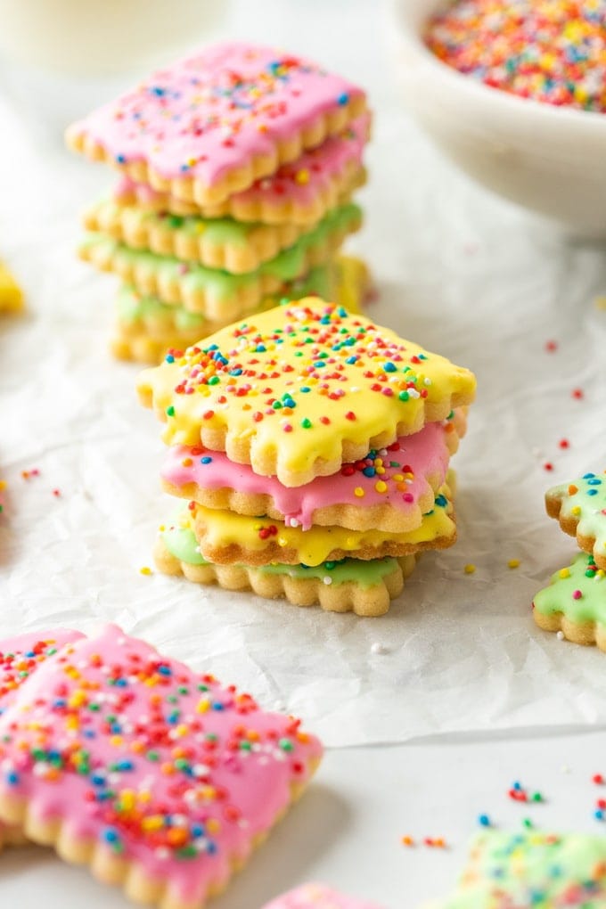 Stacks of coloured iced cookies covered in sprinkles on a sheet of baking paper.