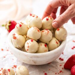 A batch of white chocolate truffles in a small white bowl with a hand reaching into pick one up