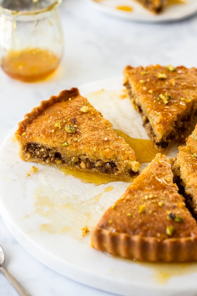 3 slices of baklava tart on a marble cake plate with a pot of syrup behind
