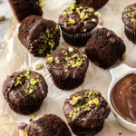 A batch of chocolate muffins on a sheet of baking paper with pistachios scattered on top and around