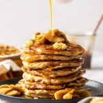A stack of pancakes topped with apples with maple syrup being drizzled over the top